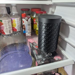 May include: A black cylindrical container with a honeycomb pattern sits inside a refrigerator. The container is on a glass shelf with other items, including bottles, jars, and food packaging. The refrigerator is white.