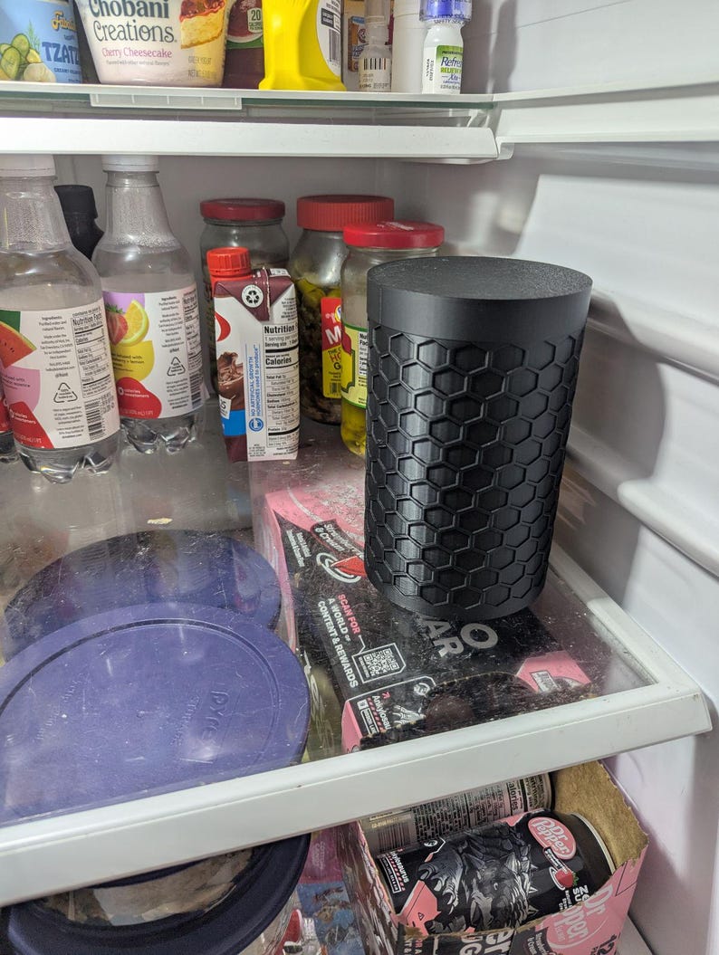 May include: A black cylindrical container with a honeycomb pattern sits inside a refrigerator. The container is on a glass shelf with other items, including bottles, jars, and food packaging. The refrigerator is white.
