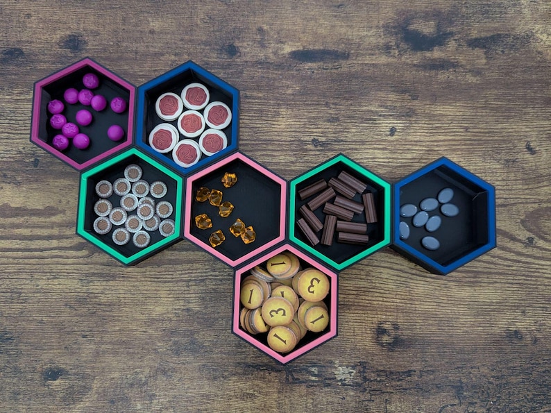 May include: A collection of black hexagon-shaped game component organizers with colorful trim, each filled with different game pieces. The pieces include purple spheres, red and white tokens, brown and white tokens, amber gems, brown rectangular pieces, and gray oval pieces.