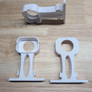 Può includere: Tre supporti per bastoni per tende in plastica bianca, ciascuno caratterizzato da una base rettangolare con un'apertura circolare per il bastone, un braccio di supporto verticale con un intaglio ovale allungato e una piastra di montaggio.