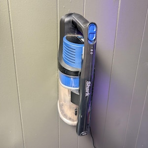 Puede incluir: Una aspiradora de mano Shark negra y azul montada en una pared gris. La aspiradora tiene un depósito de polvo transparente, un filtro azul y un asa negra con botones de control. El logotipo de Shark es visible.