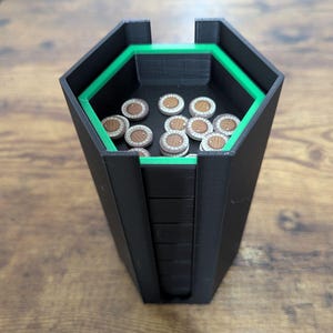 May include: Black and green hexagonal dice tower with a green interior trim. The tower holds several wooden tokens with a brown and white design. The tower is sitting on a wooden surface.