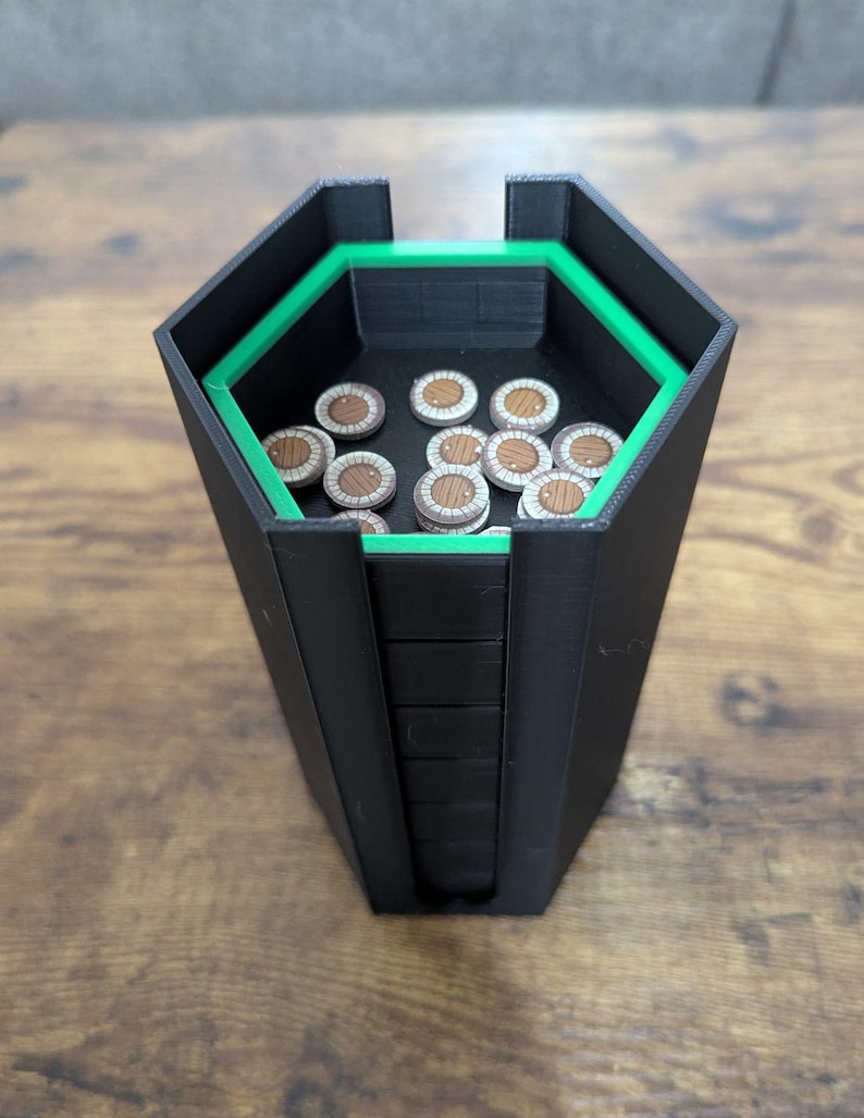 May include: Black and green hexagonal dice tower with a green interior trim. The tower holds several wooden tokens with a brown and white design. The tower is sitting on a wooden surface.