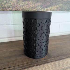 May include: A black cylindrical container with a honeycomb pattern. The container is made of a matte black material and sits on a wooden surface. The background features a landscape painting and a white brick wall.