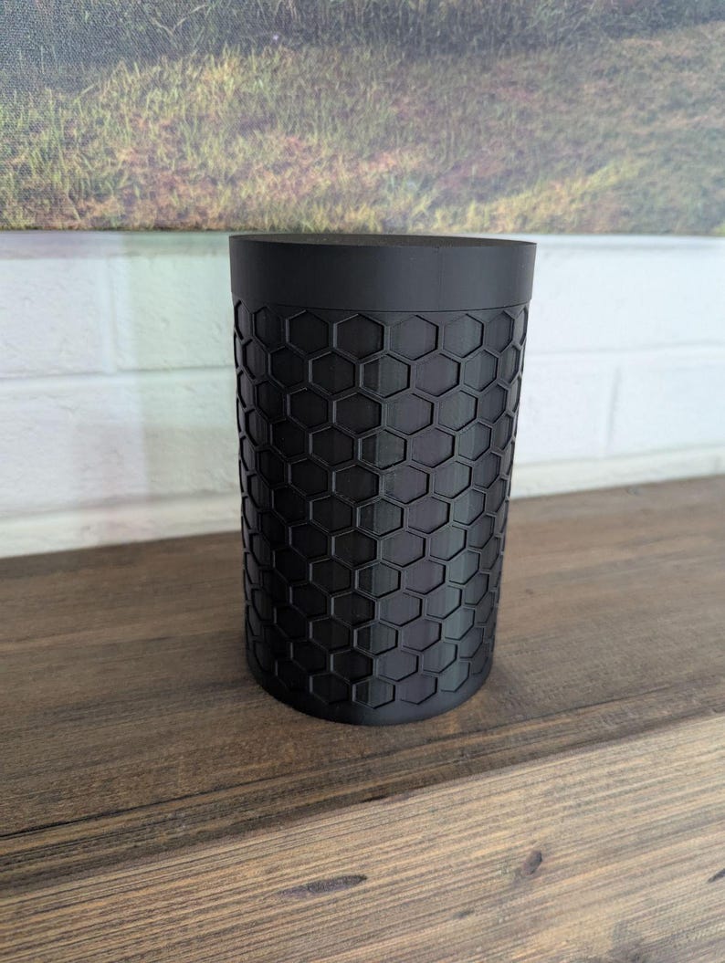 May include: A black cylindrical container with a honeycomb pattern. The container is made of a matte black material and sits on a wooden surface. The background features a landscape painting and a white brick wall.
