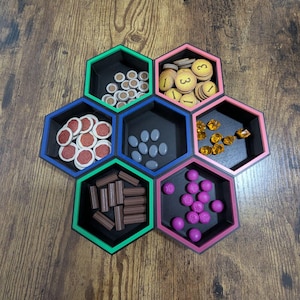 May include: A set of seven hexagonal game piece organizers in black, with colored borders of green, pink, and blue. Each hexagon holds different game pieces, including wooden tokens, gems, and dice.