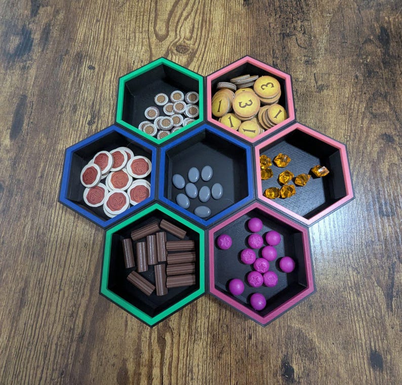 May include: A set of seven hexagonal game piece organizers in black, with colored borders of green, pink, and blue. Each hexagon holds different game pieces, including wooden tokens, gems, and dice.