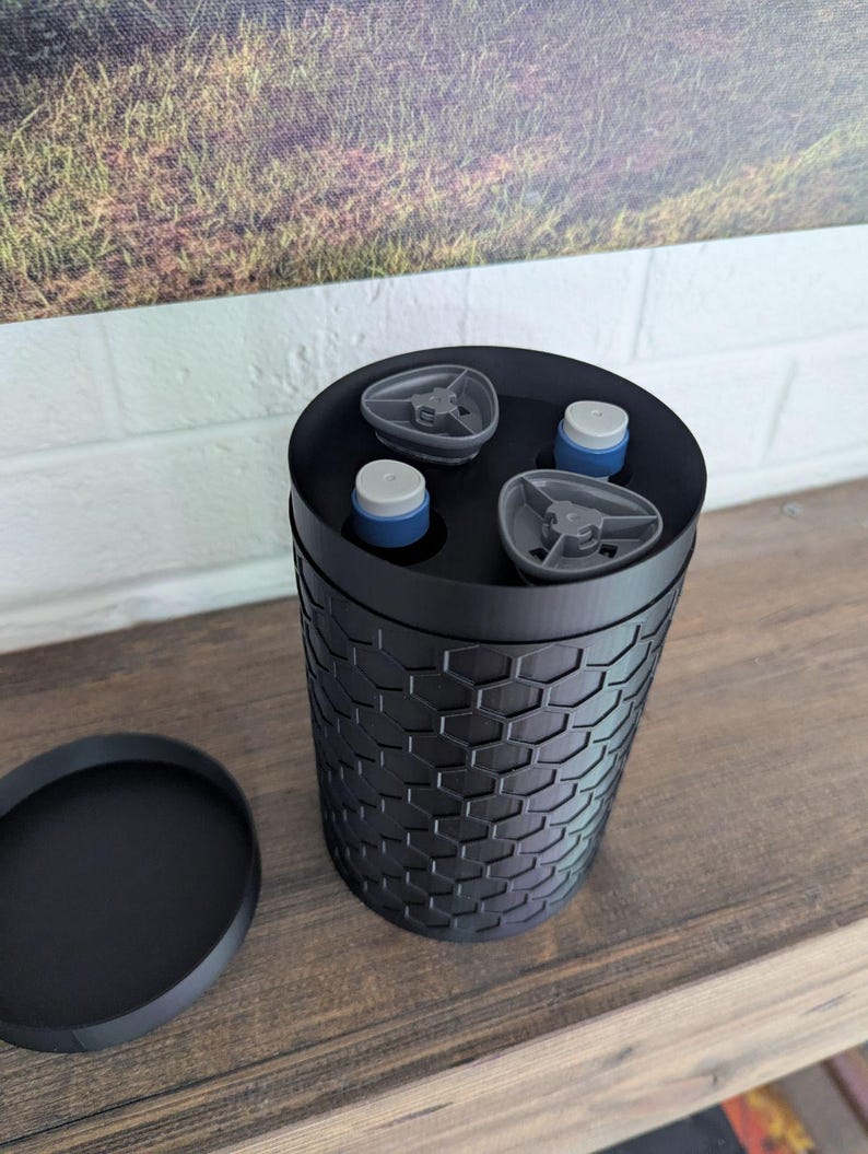 May include: A black cylindrical container with a honeycomb pattern, holding three small bottles with blue and white caps. A separate black lid sits to the side. The container is on a wooden surface.