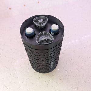 May include: A black, cylindrical container with a textured, honeycomb-like pattern. The top has three gray, triangular holders and two white and blue objects. The container is sitting on a white surface.