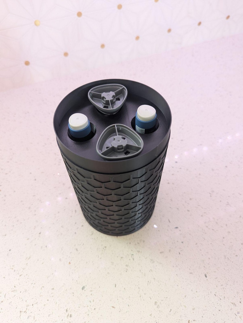 May include: A black, cylindrical container with a textured, honeycomb-like pattern. The top has three gray, triangular holders and two white and blue objects. The container is sitting on a white surface.