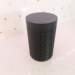 May include: A cylindrical black object with a flat top and a textured honeycomb pattern on the sides. The object is made of a solid, dark material and stands on a white surface.