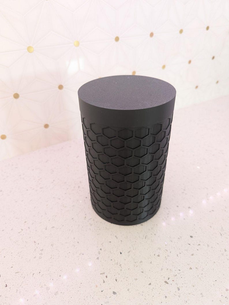May include: A cylindrical black object with a flat top and a textured honeycomb pattern on the sides. The object is made of a solid, dark material and stands on a white surface.