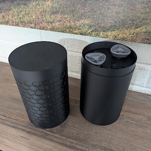 May include: Two black cylindrical containers, one open and one closed, sit on a wooden surface. The closed container has a textured, honeycomb-like pattern. The open container reveals two small, gray objects inside.