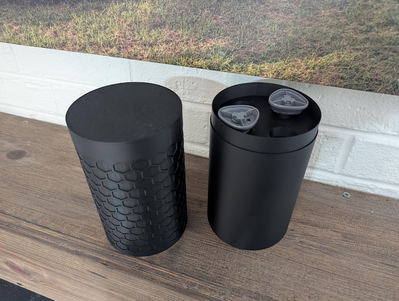 May include: Two black cylindrical containers, one open and one closed, sit on a wooden surface. The closed container has a textured, honeycomb-like pattern. The open container reveals two small, gray objects inside.