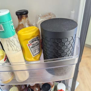 May include: A black container with a honeycomb pattern sits in a refrigerator shelf next to a bottle of yellow mustard. Other bottles of condiments are visible in the fridge.