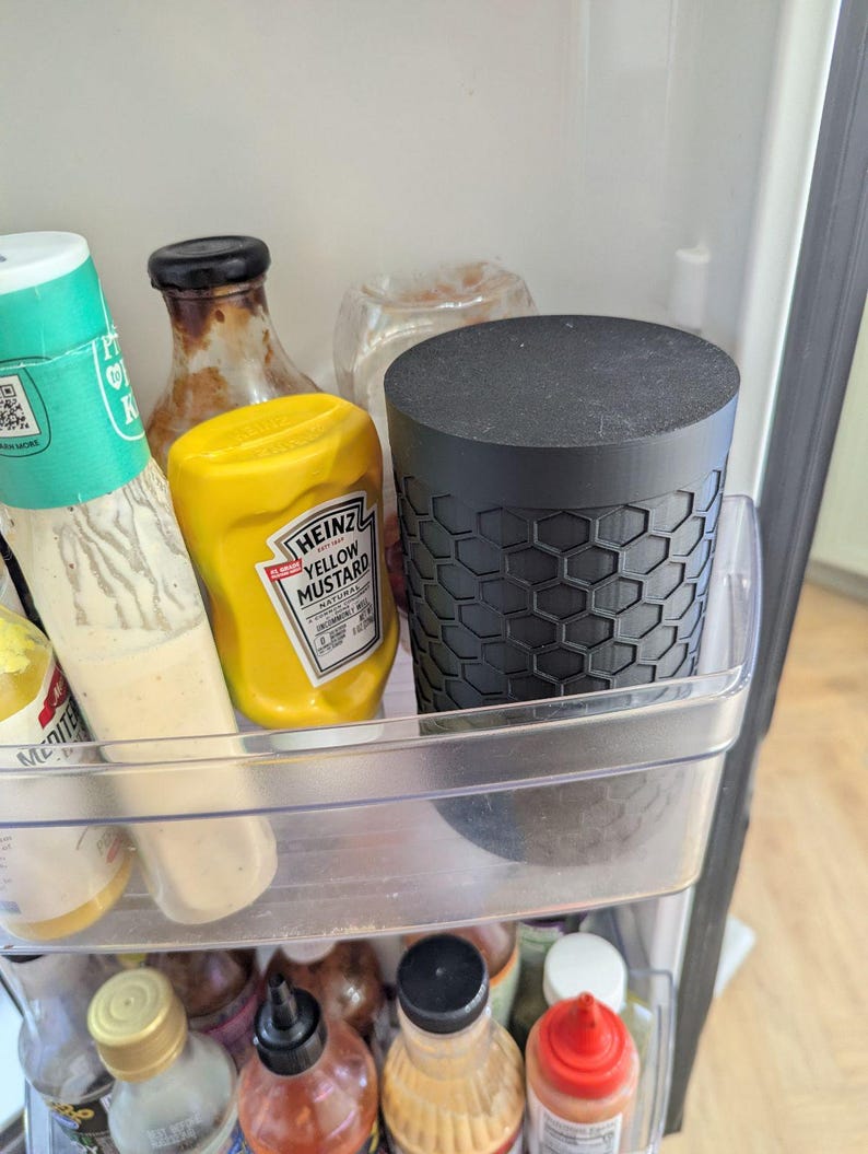 May include: A black container with a honeycomb pattern sits in a refrigerator shelf next to a bottle of yellow mustard. Other bottles of condiments are visible in the fridge.