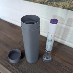 以下が含まれることがあります： 紫色のキャップが付いた白と透明の医療機器の隣にある、蓋付きの灰色の円筒形のケース。ケースは開いており、内部が明らかになっています。医療機器には側面にテキストが印刷されています。