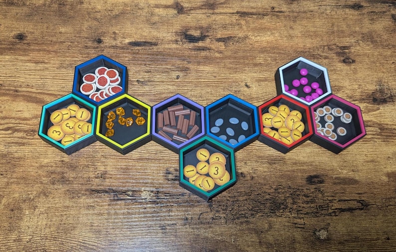 May include: A set of black hexagonal game organizers filled with various game pieces. The pieces include red and white tokens, yellow coins, brown wooden pieces, and purple spheres. The organizers are on a wooden surface.