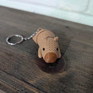 May include: A light brown capybara keyring with a silver ring. The capybara has a textured, knitted appearance with a brown nose and small black eyes. The keyring is on a dark wooden surface.