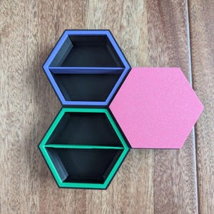 May include: Three hexagonal shelves in a honeycomb arrangement. Two shelves are black with colored trim in purple and green, each with a divider. A pink hexagon is next to the shelves. The shelves are on a wooden surface.