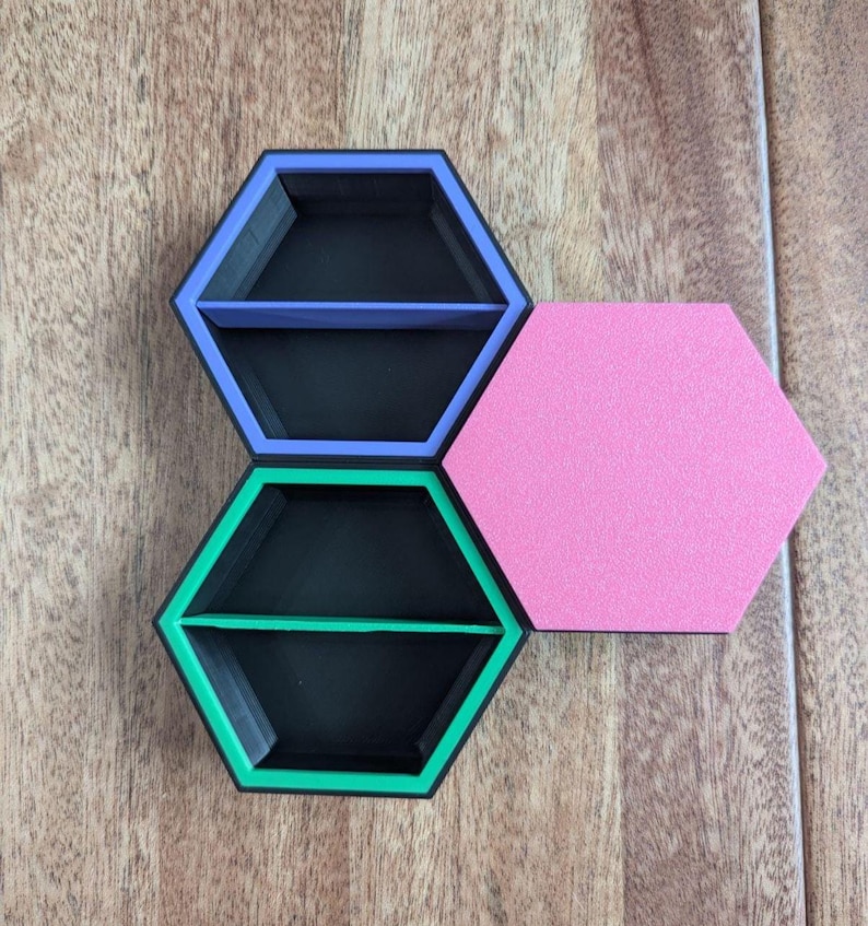 May include: Three hexagonal shelves in a honeycomb arrangement. Two shelves are black with colored trim in purple and green, each with a divider. A pink hexagon is next to the shelves. The shelves are on a wooden surface.