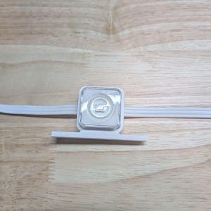 Può includere: Un modulo luminoso LED quadrato bianco con una lente centrale trasparente, montato su una base piatta in plastica bianca. La luce è integrata in un sottile cavo elettrico bianco che attraversa orizzontalmente il centro dell'alloggiamento.