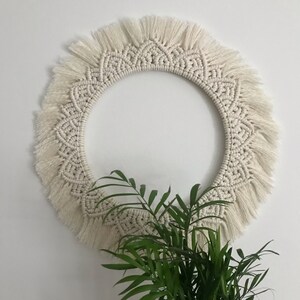 May include: A white macrame wall hanging with a circular shape and a fringe border. The hanging is made with a knotting technique and has a delicate, intricate design.