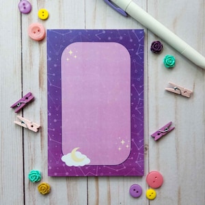 May include: A purple notepad with a galaxy design and a white border. The notepad has a pink interior with a crescent moon and stars. The notepad is surrounded by colorful buttons and paper clips.
