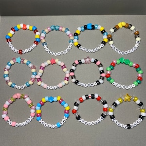May include: Twelve colorful beaded bracelets with different names on them, including "Hello Kitty", "Cinnamoroll", "Choccat", "Pompompurin", "Kiki", "Lala", "Kuromi", "Keroppi", "My Melody", "Tuxedo Sam", "Pochacco", and "Badtz Maru".