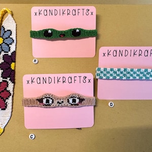 May include: Four friendship bracelets on pink cards with the text "xKANDIKRAFTSx". The bracelets are made with colorful thread and feature different designs, including flowers, a green frog face, a brown cat face, and a blue and white checkered pattern.