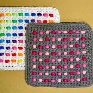 May include: Two square crochet potholders. One is white with rows of multi-colored plastic beads in shades of orange, yellow, green, blue, purple, and red. The other is gray with rows of alternating light pink and dark pink plastic beads.