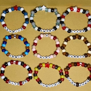 May include: Nine circular beaded bracelets arranged in a grid. Each bracelet features colorful plastic pony beads, letter beads spelling names or phrases like REMMICK, SMOKE, STACK, ANNIE, MARY, SAMMIE, BITE ME, SINNERS, and CLUB JUKE, and star or heart charms.