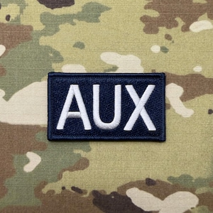 May include: A rectangular navy blue patch with the white embroidered letters "AUX". The patch is set against a camouflage background with shades of green, brown, and tan.