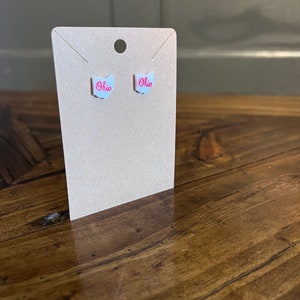 May include: A pair of white stud earrings with a pink outline of the state of Ohio and the word "Ohio" written inside. The earrings are displayed on a brown card.