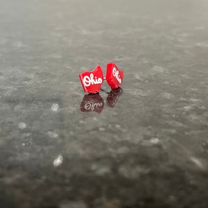 May include: A pair of red stud earrings shaped like the state of Ohio with the word "Ohio" written in white lettering.