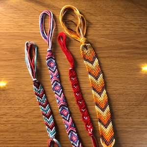 Könnte beinhalten: Vier handgefertigte, gewebte Freundschaftsarmbänder mit komplizierten Knüpfmustern. Die Armbänder zeigen verschiedene Designs wie Zickzacklinien, Rauten und Herzmotive in lebendigen Farbpaletten aus Rot, Orange, Gelb, Pink, Blau und Weiß.