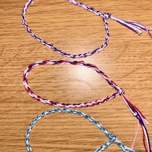 Può includere: Tre braccialetti dell'amicizia intrecciati nei toni del viola, rosso e blu. Ogni braccialetto è realizzato con più fili e presenta una chiusura ad anello e nappina. I braccialetti sono esposti su una superficie di legno.