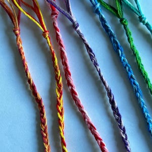 May include: Close-up of seven braided friendship bracelets in various colours, including red, yellow, orange, pink, purple, blue, and green. Each bracelet has a loop at the top for tying. The bracelets are made of colourful threads.