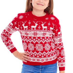 以下が含まれることがあります： 赤い白のクリスマスセーターを着た若い女の子。セーターにはトナカイの模様が描かれています。セーターはクルーネックで長袖です。女の子は青いジーンズを履いています。