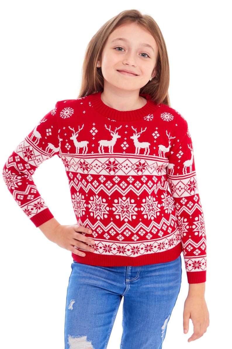 以下が含まれることがあります： 赤い白のクリスマスセーターを着た若い女の子。セーターにはトナカイの模様が描かれています。セーターはクルーネックで長袖です。女の子は青いジーンズを履いています。
