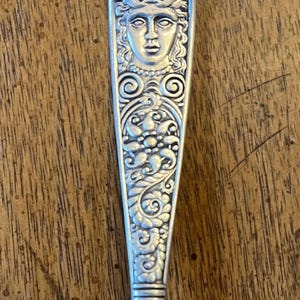 May include: A silver spoon with an ornate handle featuring a woman's face with a crown and floral designs.