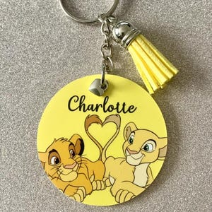 May include: Yellow keychain featuring a cartoon lion and lioness. The keychain has a yellow tassel and a silver-coloured metal ring. The name "Charlotte" is written in black script on the yellow background.