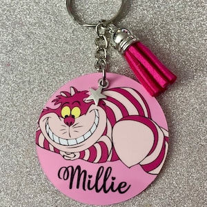 May include: A pink round keychain featuring the Cheshire Cat from Alice in Wonderland. The cat has a wide grin and striped pink and white fur. The keychain includes a silver chain, a pink tassel, and a star charm. The name "Millie" is written on the keychain.