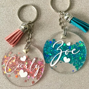 May include: Two personalised keyrings with clear, round pendants filled with glitter. One has pink glitter and the name "Carly" in white script, the other has teal glitter and the name "Zoe". Both have heart accents and matching tassels in pink and teal.