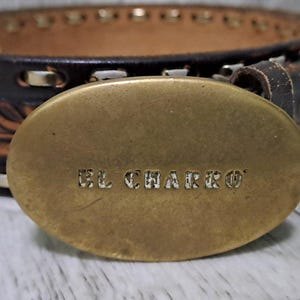 May include: A dark brown leather belt featuring an oval-shaped brass buckle with the text EL CHARRO embossed in the center. The belt strap displays a decorative floral tooling pattern along the edges and is accented with silver-toned metal stitching.