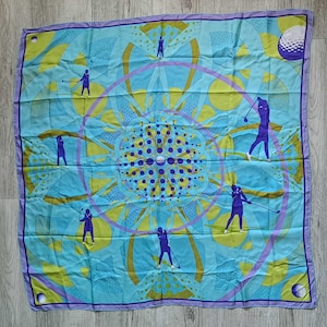 May include: A square silk scarf featuring a light blue and lime green geometric pattern with a central mandala design. The scarf is decorated with repeating silhouettes of golfers in purple, along with purple golf ball motifs in the corners.