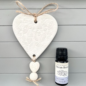 May include: White ceramic heart-shaped diffuser with a floral design, hanging from a twine string. Three white ceramic beads hang from the bottom of the heart. A small bottle of essential oil labelled "Ocean Breeze" is next to the diffuser.