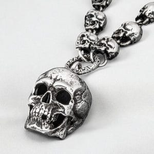 May include: A silver-colored necklace featuring a large skull pendant and a chain of smaller skull links. The necklace is displayed on a white surface, highlighting the intricate details of the skull design.