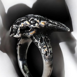 May include: A silver ring featuring a detailed, textured design of a creature's head. The ring is made of metal and has a wide band.