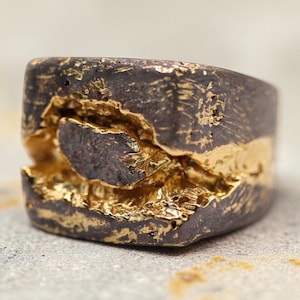 May include: A ring featuring a dark, oxidized metal band with a rough, textured surface. A horizontal fissure runs across the center of the band, revealing a bright, polished gold-colored interior that contrasts with the dark exterior finish.